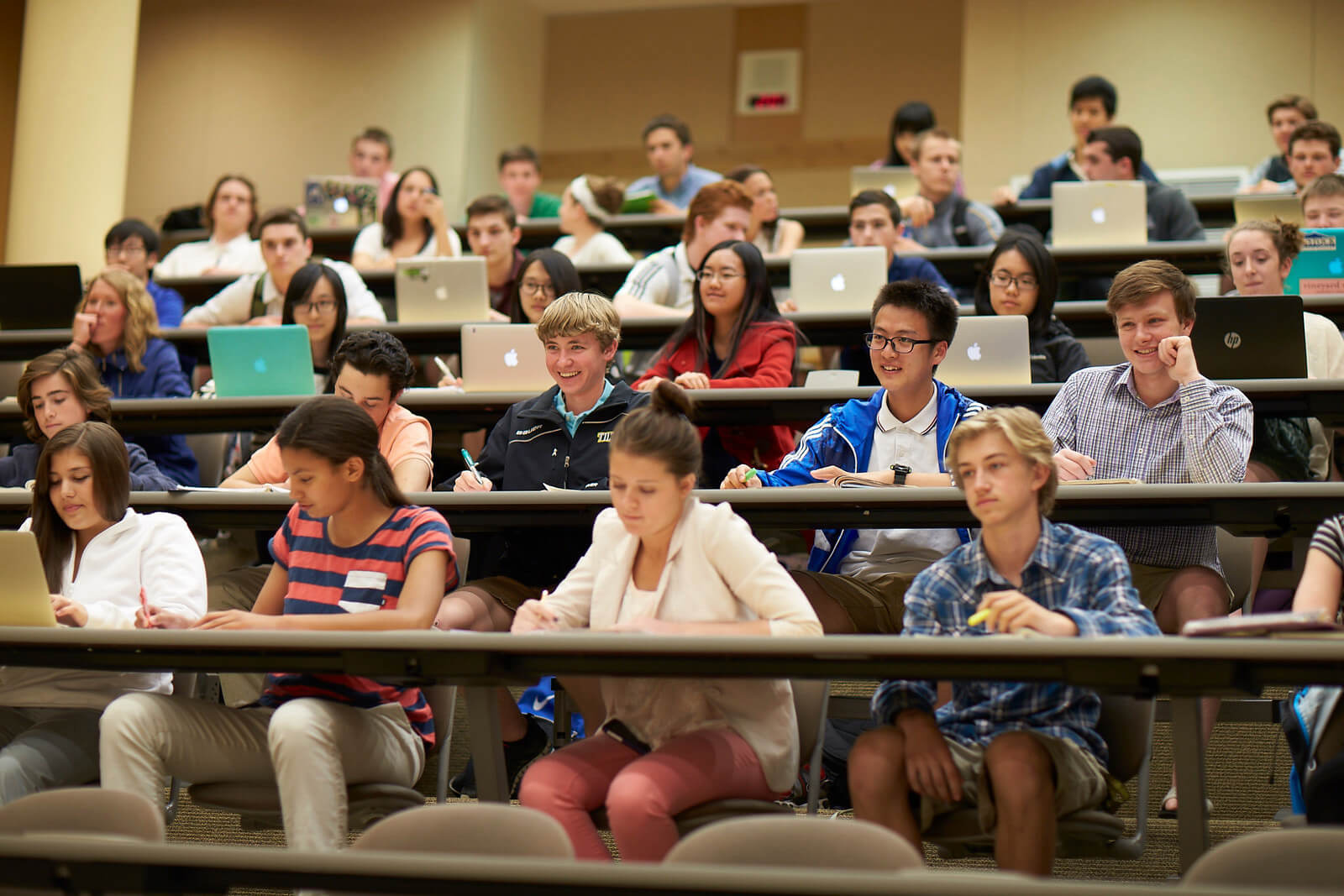 Aula con gradas, con muchos estudiantes internacionales cogiendo apuntes y con portátiles Apple y HP