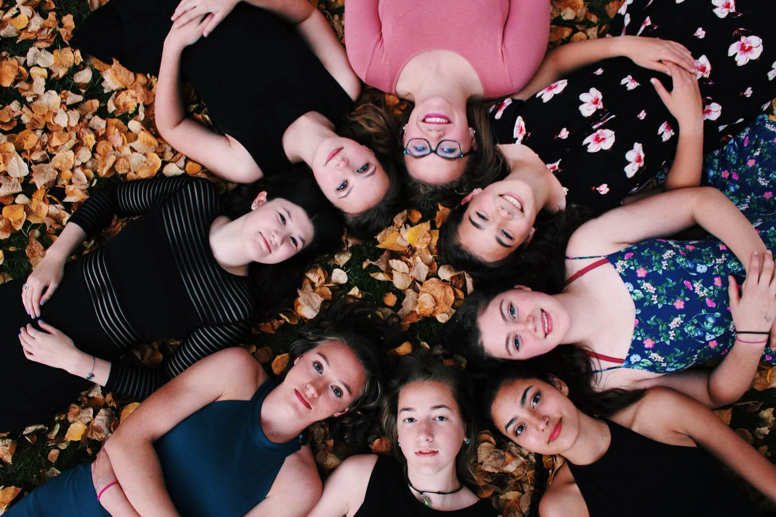 Ocho amigas tendidas en el suelo sobre hojas de otoño, fotografiadas desde arriba.